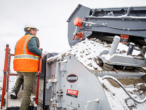 A worker in a hard hat and safety vest inspects the open rotor chamber of a Superior Sentry horizontal shaft impact crusher at a snowy job site.