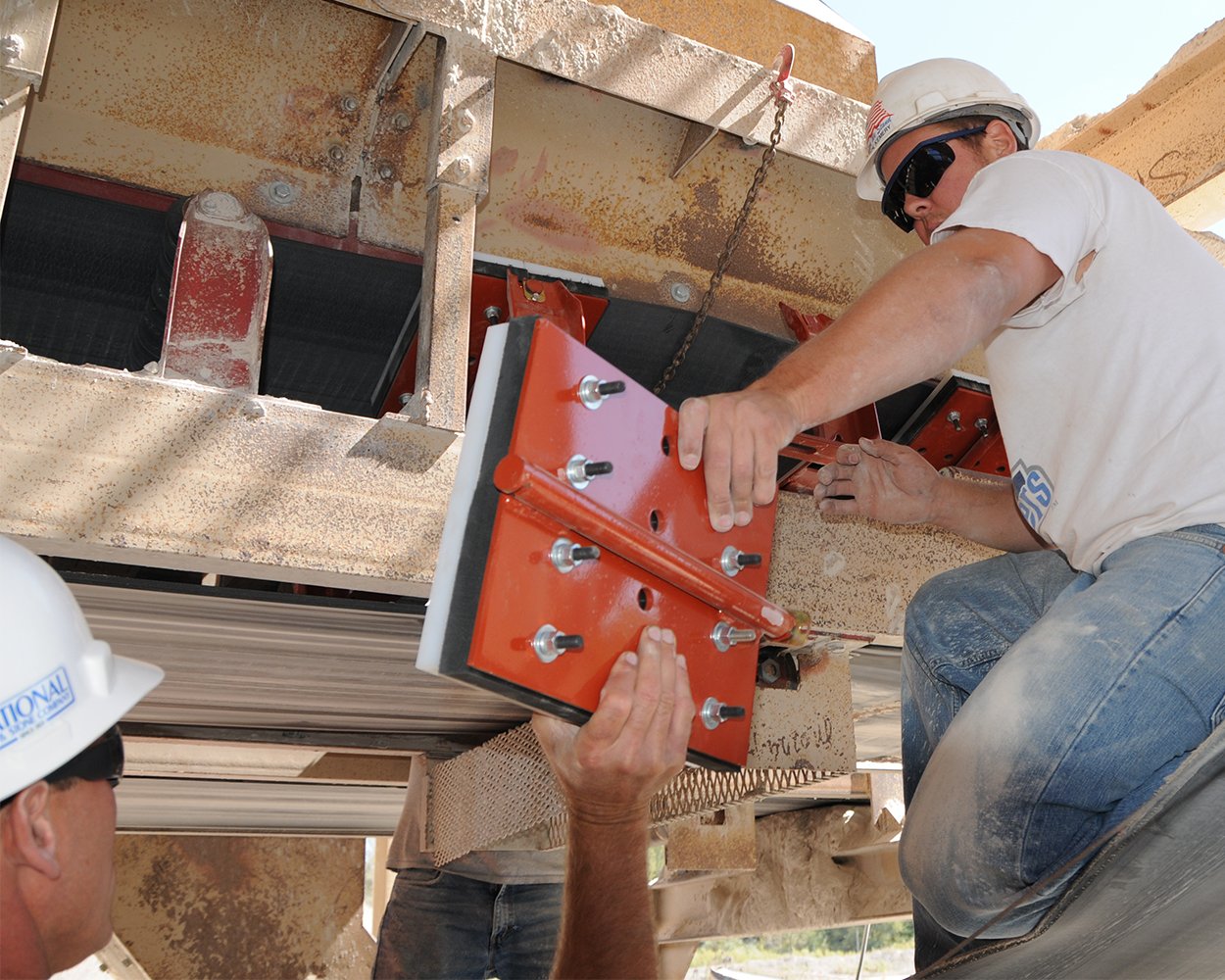 Technicians installing a conveyor belt sealing system cartridge on an idler frame beneath a load zone to support the belt and prevent spillage