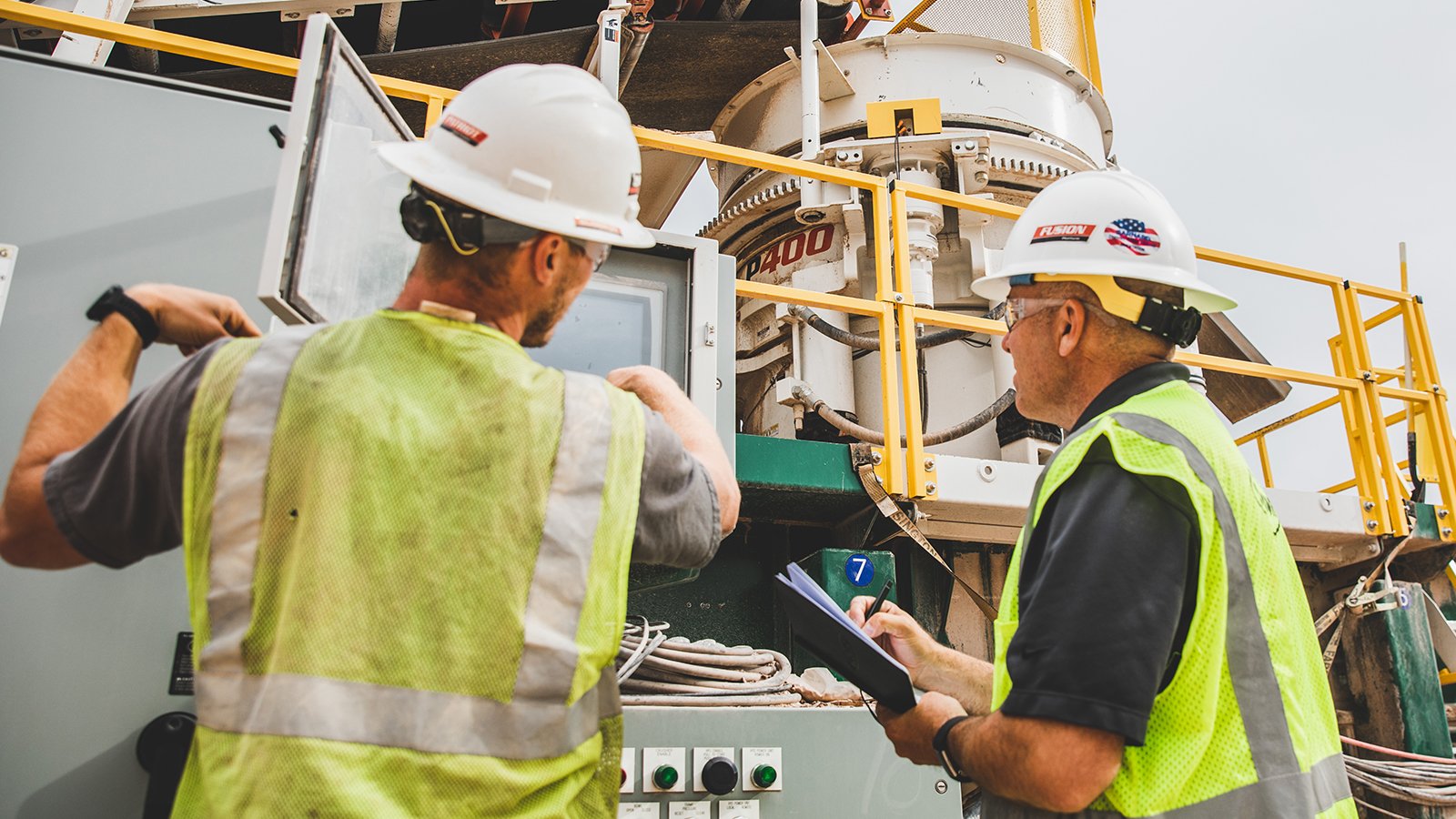 Two quarry workers in safety vests and hard hats review a control panel and take notes beside an automated cone crusher.