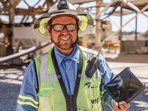 Quarry worker in safety gear uses a tablet to control plant operations, with a radio clipped to his vest at an aggregate site.
