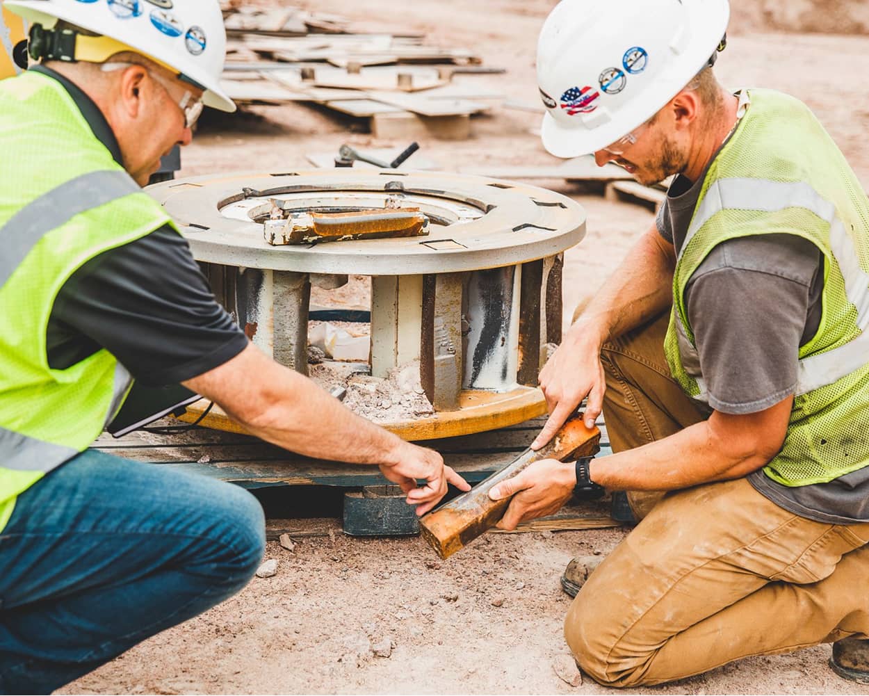 Two workers in safety vests and hard hats inspect and install wear shoes on a VSI crusher rotor.
