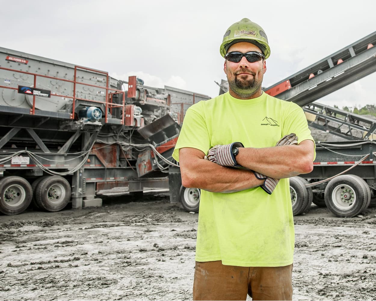 Worker wearing a hard hat and safety glasses stands in front of portable screening equipment at a quarry site