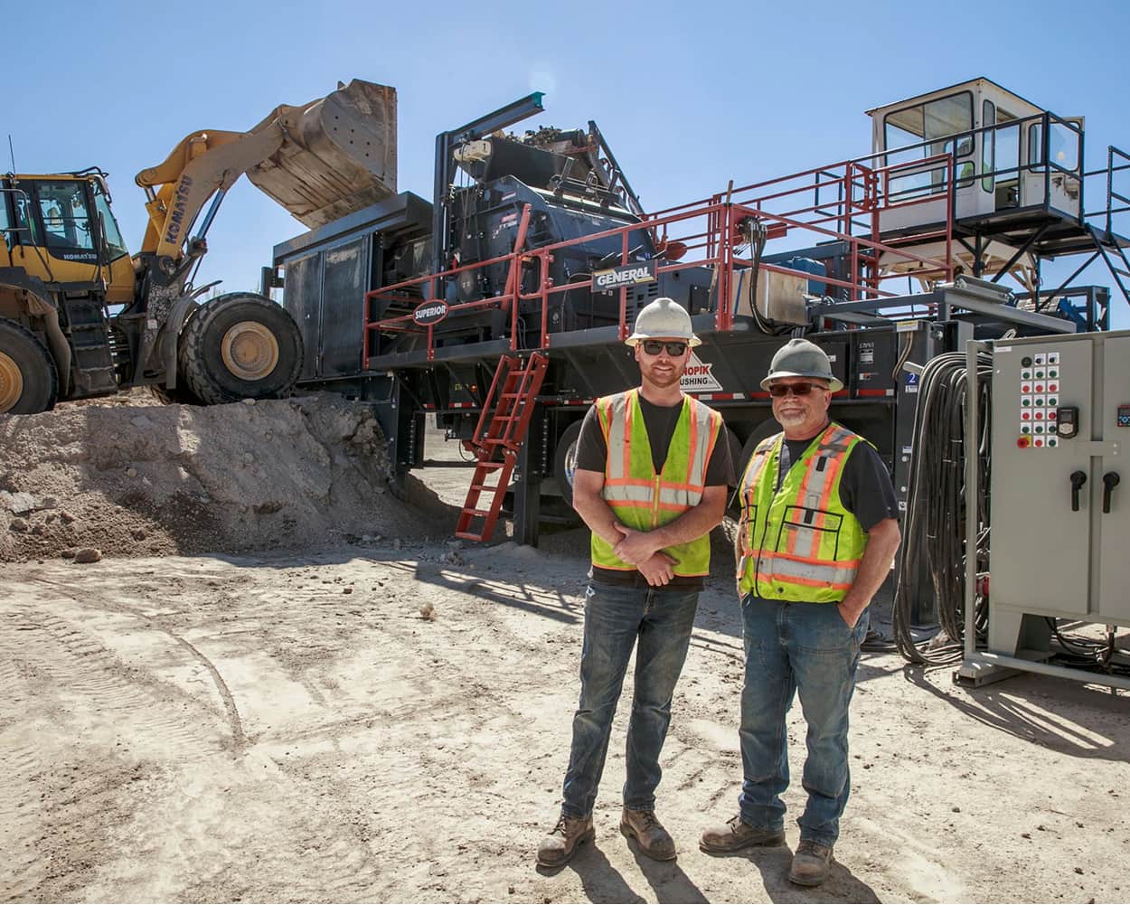 Two workers in safety vests standing in front of a Superior Industries portable crushing plant as a loader feeds material into the hopper at an active job site
