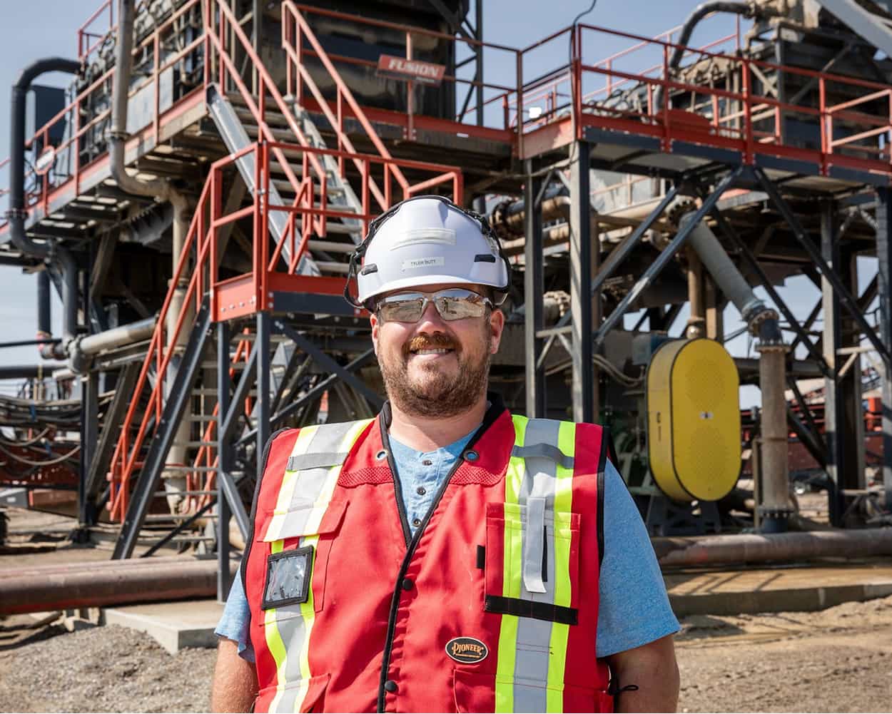 Worker wearing a hard hat and safety vest stands in front of the Fusion Wash Plant with a classifying tank in Alberta