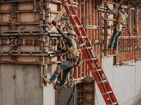 Two construction workers wearing safety gear and harnessing secure formwork on a concrete structure during the construction and installation phase of a large industrial project