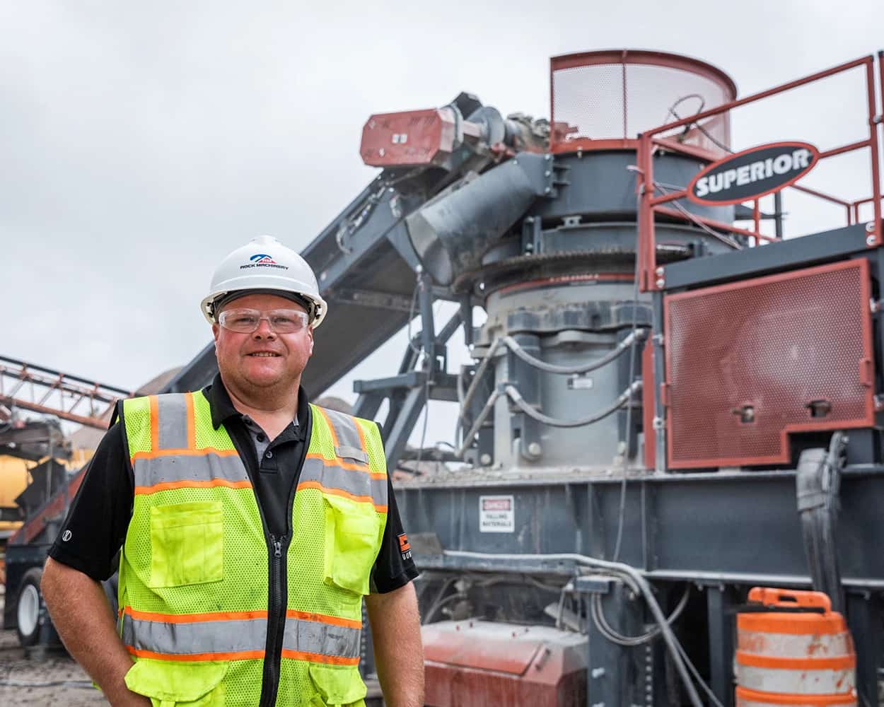 Operator in hard hat and safety vest standing in front of paused Patriot Cone Crusher operation.