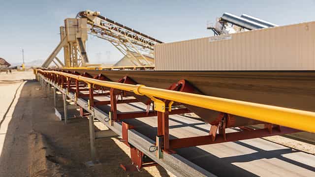 Conveyor system with a yellow handrail installed for worker safety, running alongside a belt carrying bulk material at an industrial site.