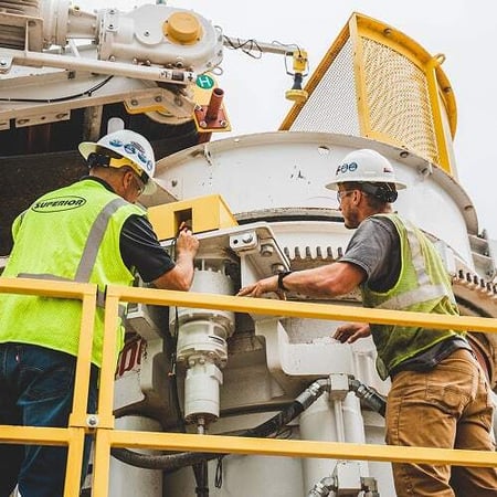 Two workers in safety vests and hard hats inspect and service a large cone crusher at a quarry site in Colorado